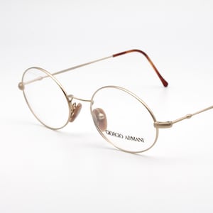 May include: Gold-tone Giorgio Armani eyeglasses with oval lenses. The frames have a thin, delicate design with a brown accent on the temple tips. The brand name is printed on the lens.