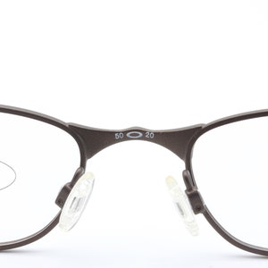 May include: A pair of brown metal spectacles with clear lenses. The glasses have a small oval logo on the bridge of the nose with the text "50 20".