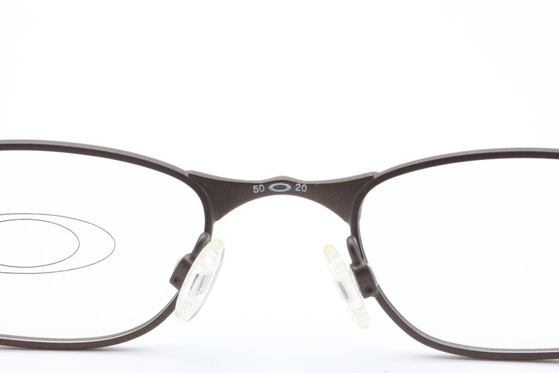 May include: A pair of brown metal spectacles with clear lenses. The glasses have a small oval logo on the bridge of the nose with the text "50 20".