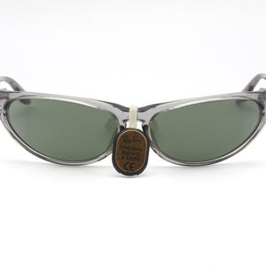 May include: Gray translucent sunglasses with green lenses. The sunglasses have a curved, cat-eye shape. A small brown tag with the text "ORIGINAL BAUSCH & LOMB" is attached to the sunglasses.