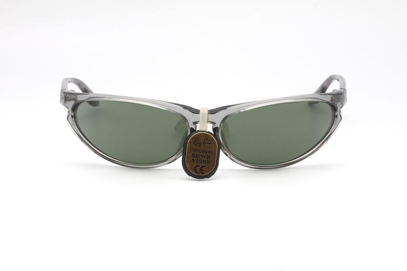 May include: Gray translucent sunglasses with green lenses. The sunglasses have a curved, cat-eye shape. A small brown tag with the text "ORIGINAL BAUSCH & LOMB" is attached to the sunglasses.