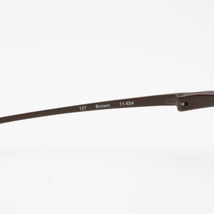 Op de afbeelding: Close-up van een bruin metalen montuur. De tekst "127 Brown 11-634" staat op de poot. Het montuur heeft een strak, modern design met een donkerbruine afwerking. De glazen zijn helder.