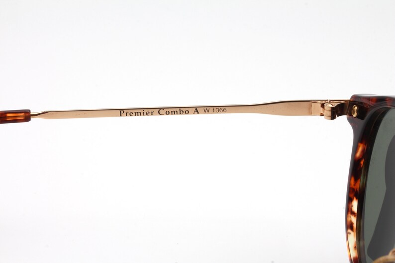 Op de afbeelding: Close-up van de gouden metalen poot van een paar schildpadzonnebrillen. De poot is gegraveerd met de tekst "Premier Combo A W 1366".