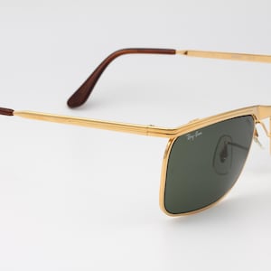 Op de afbeelding: Goudkleurige zonnebril met donkergroene glazen. De zonnebril heeft een rechthoekige vorm en bruine pootjes. Het Ray-Ban-logo is zichtbaar op het glas. De bril staat tegen een witte achtergrond.