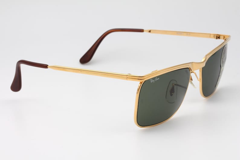 Op de afbeelding: Goudkleurige zonnebril met donkergroene glazen. De zonnebril heeft een rechthoekige vorm en bruine pootjes. Het Ray-Ban-logo is zichtbaar op het glas. De bril staat tegen een witte achtergrond.