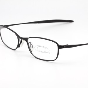 Op de afbeelding: Zwarte rechthoekige bril met een dun montuur. De bril heeft heldere glazen en een matte afwerking. Het Oakley-logo is zichtbaar op de lens. De pootjes van de bril zijn dun en licht gebogen.