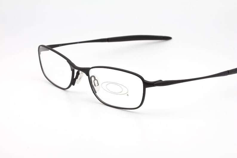 Op de afbeelding: Zwarte rechthoekige bril met een dun montuur. De bril heeft heldere glazen en een matte afwerking. Het Oakley-logo is zichtbaar op de lens. De pootjes van de bril zijn dun en licht gebogen.