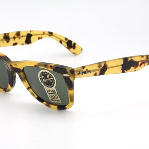 Puede incluir: Gafas de sol Ray-Ban con dise&ntilde;o Wayfarer. Las monturas presentan un patr&oacute;n de carey en tonos amarillos y marrones. Las lentes son de color verde oscuro. El logotipo de Ray-Ban es visible en la patilla y la lente.