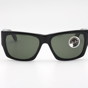 May include: Black sunglasses with a thick frame and green lenses. The sunglasses have a rectangular shape and a sticker on the right lens. The sunglasses are on a white background.