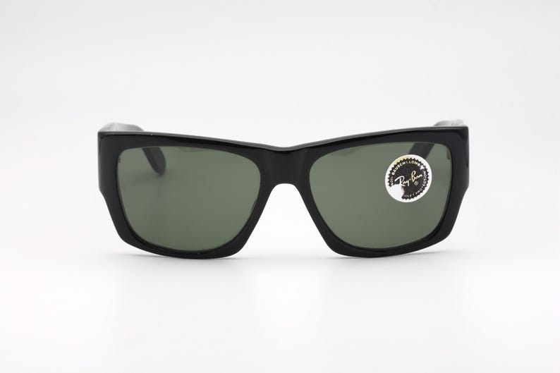 May include: Black sunglasses with a thick frame and green lenses. The sunglasses have a rectangular shape and a sticker on the right lens. The sunglasses are on a white background.