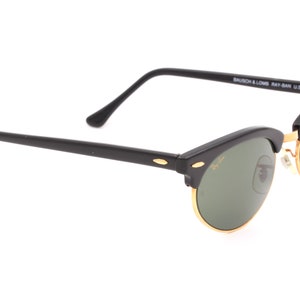 May include: Black Ray-Ban sunglasses with green lenses and gold accents. The sunglasses have a classic aviator style with a thin metal frame and a wide, rounded lens shape. The arms of the sunglasses are black and have the Ray-Ban logo on them. The sunglasses are made in the USA.