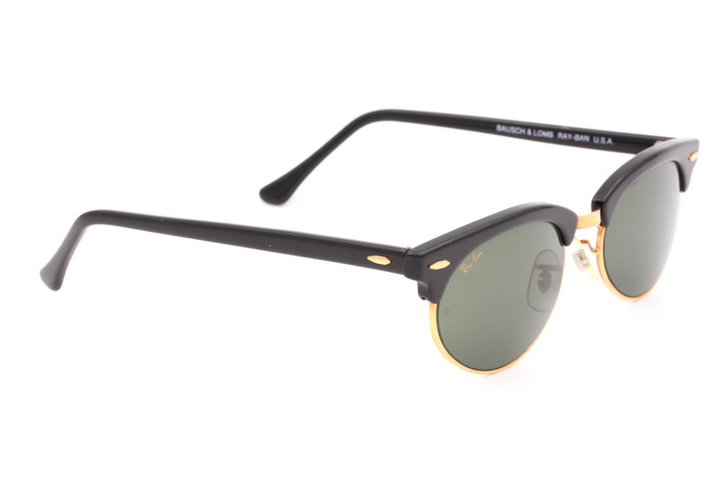 May include: Black Ray-Ban sunglasses with green lenses and gold accents. The sunglasses have a classic aviator style with a thin metal frame and a wide, rounded lens shape. The arms of the sunglasses are black and have the Ray-Ban logo on them. The sunglasses are made in the USA.