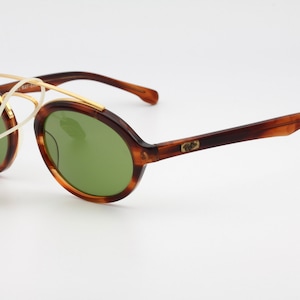 May include: Brown tortoise shell sunglasses with gold accents and green lenses. The sunglasses have a round shape and a thin gold metal bridge. The brand name is visible on the right temple.