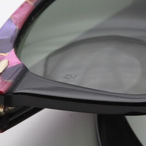 May include: Close-up of a pair of black sunglasses with a colorful, patterned frame. The sunglasses have a gold metal hinge and the lenses are tinted green. The text "Ray-Ban" is visible on the lens.