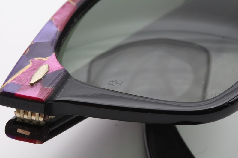 May include: Close-up of a pair of black sunglasses with a colorful, patterned frame. The sunglasses have a gold metal hinge and the lenses are tinted green. The text "Ray-Ban" is visible on the lens.