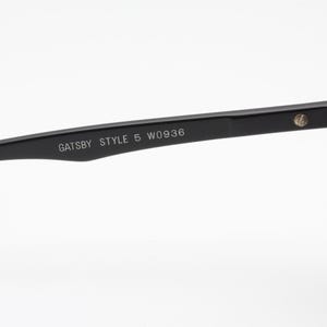 May include: Close-up of black sunglasses with the text "GATSBY STYLE 5 W0936" printed on the arm. The sunglasses have a classic design with a rectangular frame and dark lenses. The hinge and arm are black.