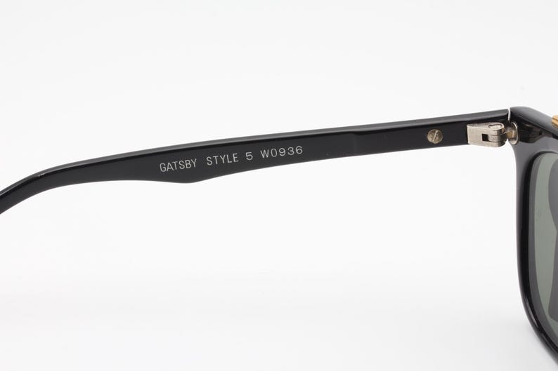 May include: Close-up of black sunglasses with the text "GATSBY STYLE 5 W0936" printed on the arm. The sunglasses have a classic design with a rectangular frame and dark lenses. The hinge and arm are black.