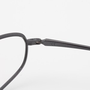 May include: Close-up of a pair of eyeglasses with a matte black finish. The frame has a rounded oval shape and a thin, angular arm. The hinge and arm are connected with small screws. The glasses are set against a white background.