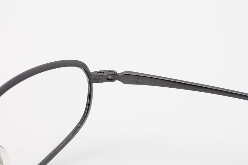 May include: Close-up of a pair of eyeglasses with a matte black finish. The frame has a rounded oval shape and a thin, angular arm. The hinge and arm are connected with small screws. The glasses are set against a white background.