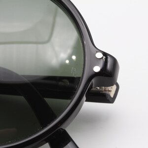 May include: Black round sunglasses with dark green lenses and a black frame. The frame has a hinge with two small white dots on the side.