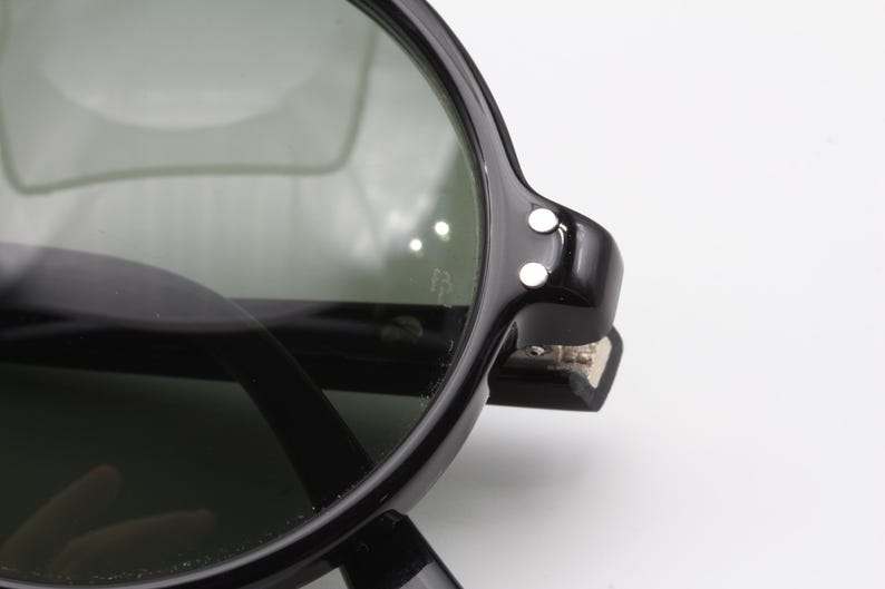 May include: Black round sunglasses with dark green lenses and a black frame. The frame has a hinge with two small white dots on the side.