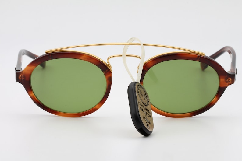 May include: Tortoise shell frame sunglasses with green lenses and a gold metal bridge. The sunglasses have a small tag attached to the frame that reads "Original Bausch & Lomb".
