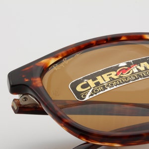 May include: Close-up of Ray-Ban sunglasses with a tortoiseshell frame and brown lenses. The sunglasses have a sticker that reads "CHROMX COLOR CONTRAST TECHNOLOGY". The Ray-Ban logo is visible on the lens.