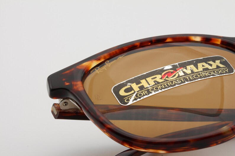 May include: Close-up of Ray-Ban sunglasses with a tortoiseshell frame and brown lenses. The sunglasses have a sticker that reads "CHROMX COLOR CONTRAST TECHNOLOGY". The Ray-Ban logo is visible on the lens.