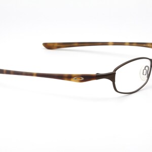May include: Brown metal-framed eyeglasses with clear lenses. The temples have a tortoise shell pattern. The glasses have a minimalist design and are set against a white background.