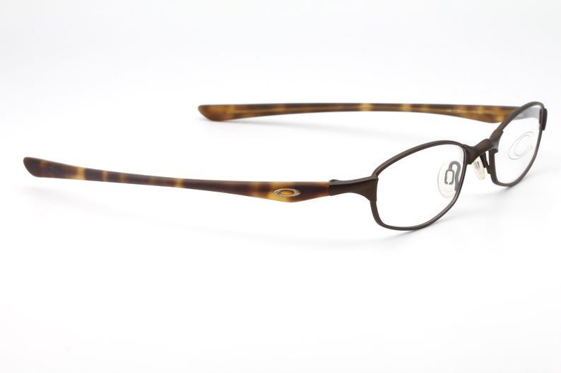 May include: Brown metal-framed eyeglasses with clear lenses. The temples have a tortoise shell pattern. The glasses have a minimalist design and are set against a white background.