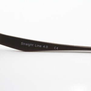 May include: Close-up of the arm of a pair of spectacles. The arm is made of dark metal and has the text "Straight Line 4.0 CE" printed on it.