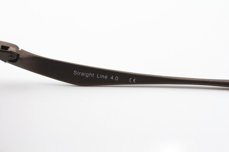 May include: Close-up of the arm of a pair of spectacles. The arm is made of dark metal and has the text "Straight Line 4.0 CE" printed on it.