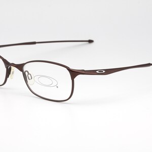 May include: Brown metal frame eyeglasses with clear lenses. The frame has a thin, curved design and a small oval logo on the side.