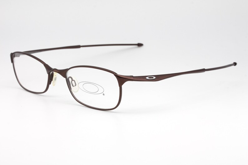 May include: Brown metal frame eyeglasses with clear lenses. The frame has a thin, curved design and a small oval logo on the side.