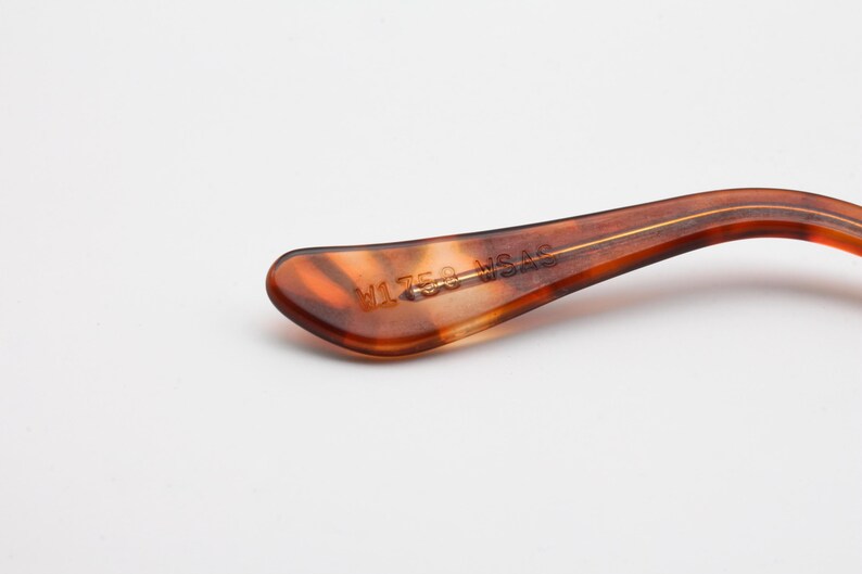 May include: Close-up of a brown and amber-colored eyeglass arm. The arm has a curved shape with a gold-colored stripe running down the center. The text "V1758 WSAS" is printed on the arm.