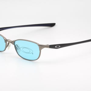 May include: A pair of Oakley eyeglasses with a silver frame and blue lenses. The temples are black with the Oakley logo. The glasses are set against a white background.