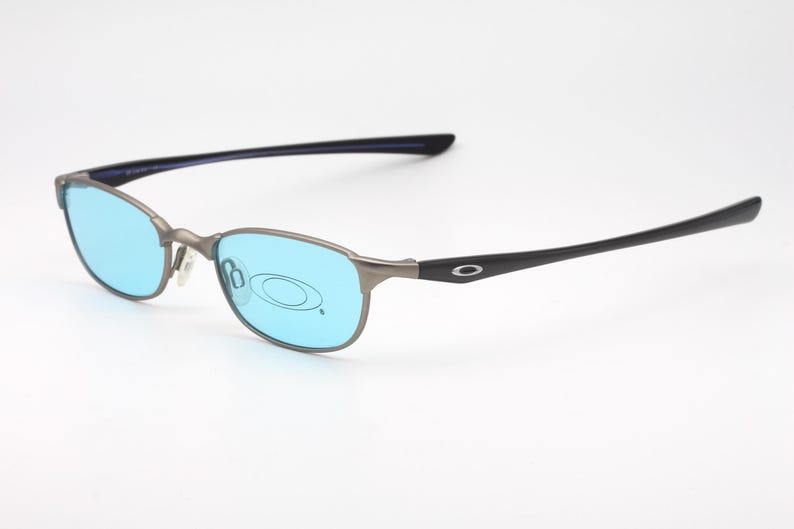 May include: A pair of Oakley eyeglasses with a silver frame and blue lenses. The temples are black with the Oakley logo. The glasses are set against a white background.