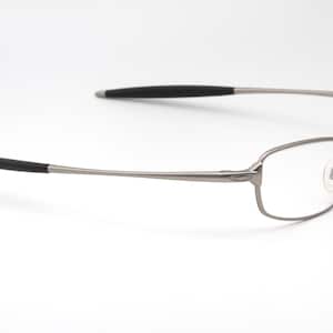 May include: A pair of eyeglasses with a rectangular frame and clear lenses. The frame is a muted silver color, with black accents on the temples. The glasses are designed for everyday use.
