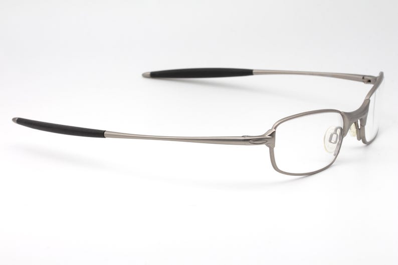 May include: A pair of eyeglasses with a rectangular frame and clear lenses. The frame is a muted silver color, with black accents on the temples. The glasses are designed for everyday use.