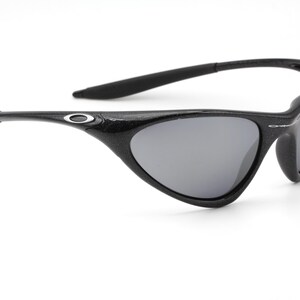 Puede incluir: Gafas de sol negras con lentes grises. Las gafas tienen una montura y patillas curvas, con un peque&ntilde;o logotipo plateado en las patillas. Las lentes son de color gris oscuro y la montura parece estar hecha de un material texturizado.