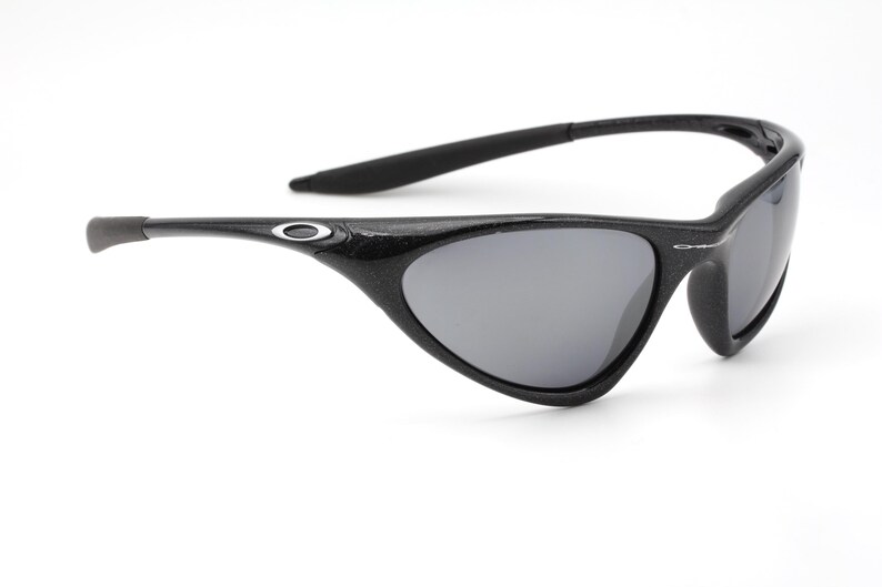 Puede incluir: Gafas de sol negras con lentes grises. Las gafas tienen una montura y patillas curvas, con un peque&ntilde;o logotipo plateado en las patillas. Las lentes son de color gris oscuro y la montura parece estar hecha de un material texturizado.
