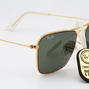 May include: Gold-framed Ray-Ban sunglasses with green lenses. The sunglasses have a classic aviator style with a square frame. The brand name "Ray-Ban" is visible on the frame. The sunglasses are accompanied by a black and gold tag that reads "Original Bausch & Lomb".