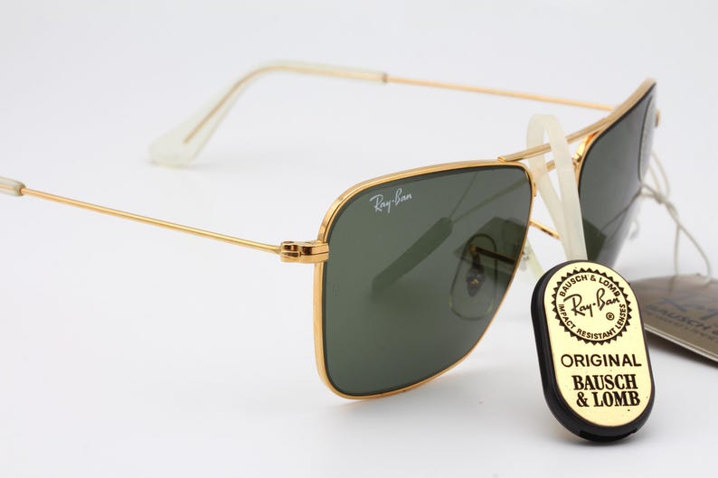May include: Gold-framed Ray-Ban sunglasses with green lenses. The sunglasses have a classic aviator style with a square frame. The brand name "Ray-Ban" is visible on the frame. The sunglasses are accompanied by a black and gold tag that reads "Original Bausch & Lomb".