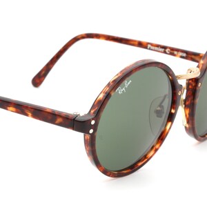 May include: Tortoiseshell Ray-Ban sunglasses with round green lenses and gold accents. The sunglasses have the text "Ray-Ban" and "Premier C" on the frame.