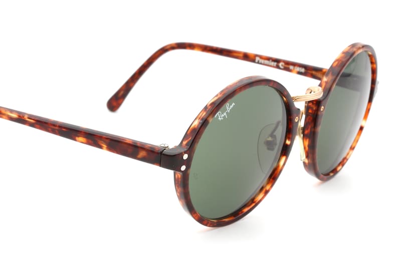 May include: Tortoiseshell Ray-Ban sunglasses with round green lenses and gold accents. The sunglasses have the text "Ray-Ban" and "Premier C" on the frame.