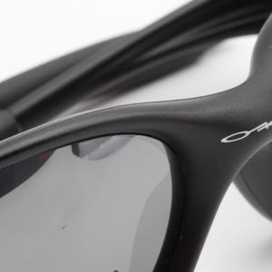 May include: Close-up of black sunglasses with gray lenses. The sunglasses have a curved design and the brand name "Oakley" is visible in white on the frame. The temples are also black and curved.