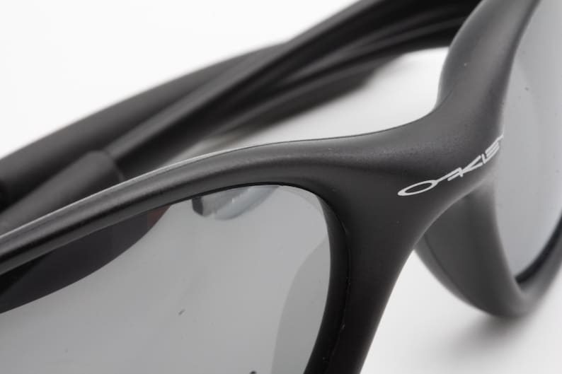 May include: Close-up of black sunglasses with gray lenses. The sunglasses have a curved design and the brand name "Oakley" is visible in white on the frame. The temples are also black and curved.