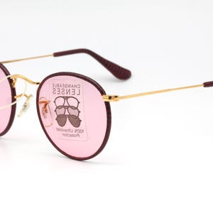May include: Gold-rimmed round sunglasses with pink lenses and burgundy frames. The lenses have a label that reads "CHANGEABLE LENSES 100% Ultraviolet Protection".