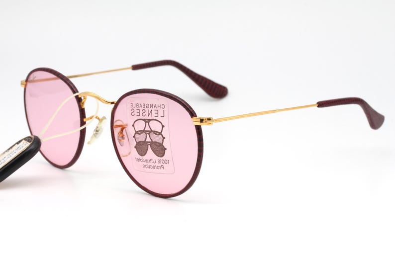 May include: Gold-rimmed round sunglasses with pink lenses and burgundy frames. The lenses have a label that reads "CHANGEABLE LENSES 100% Ultraviolet Protection".