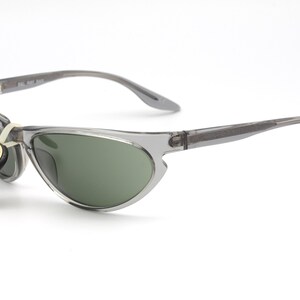 May include: Gray translucent sunglasses with green lenses. The sunglasses have a thin, curved frame and a small tag attached to the left arm that reads "Ray-Ban Original Bausch & Lomb CE".